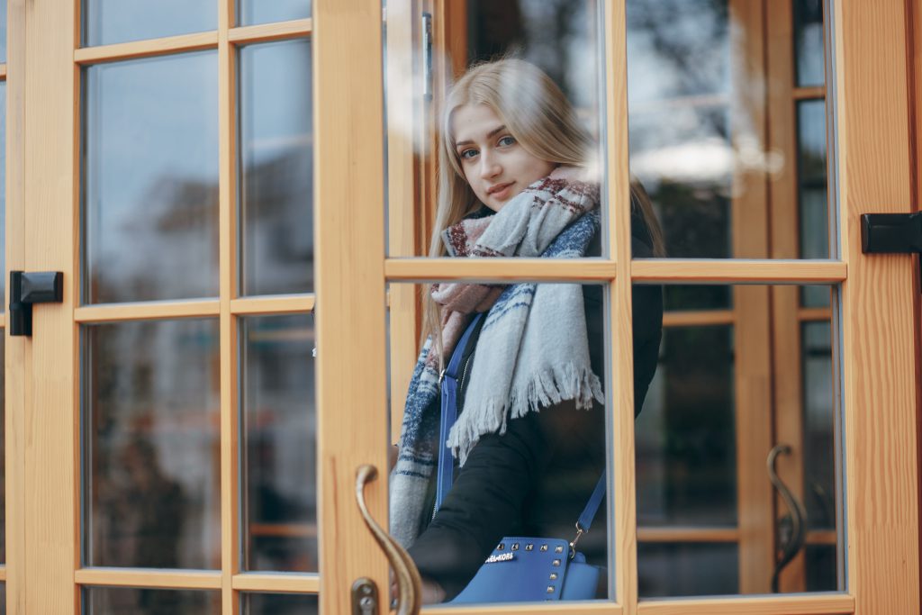Portret młodej kobiety w jasnym szaliku, patrzącej przez przeszklone okna drewniane o jasnej, naturalnej barwie, trzymającej dłoń na ozdobnej klamce.